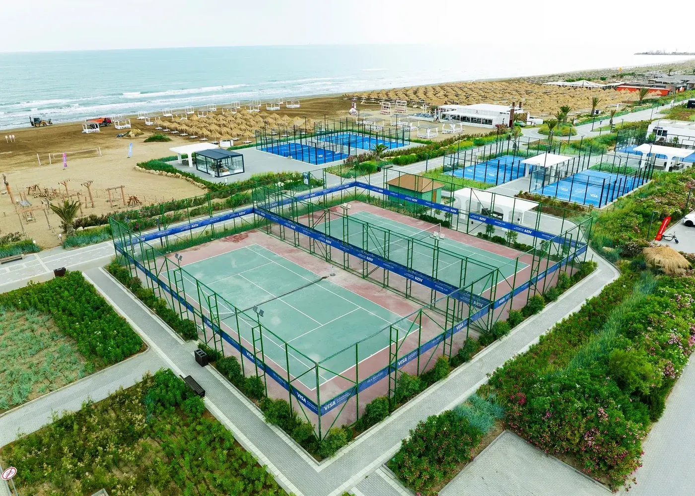 Çimərlikdə idman zonası yuxarıdan: yaşıl tennis kortunun ətrafında mavi padel kortları, arxada qum və dəniz