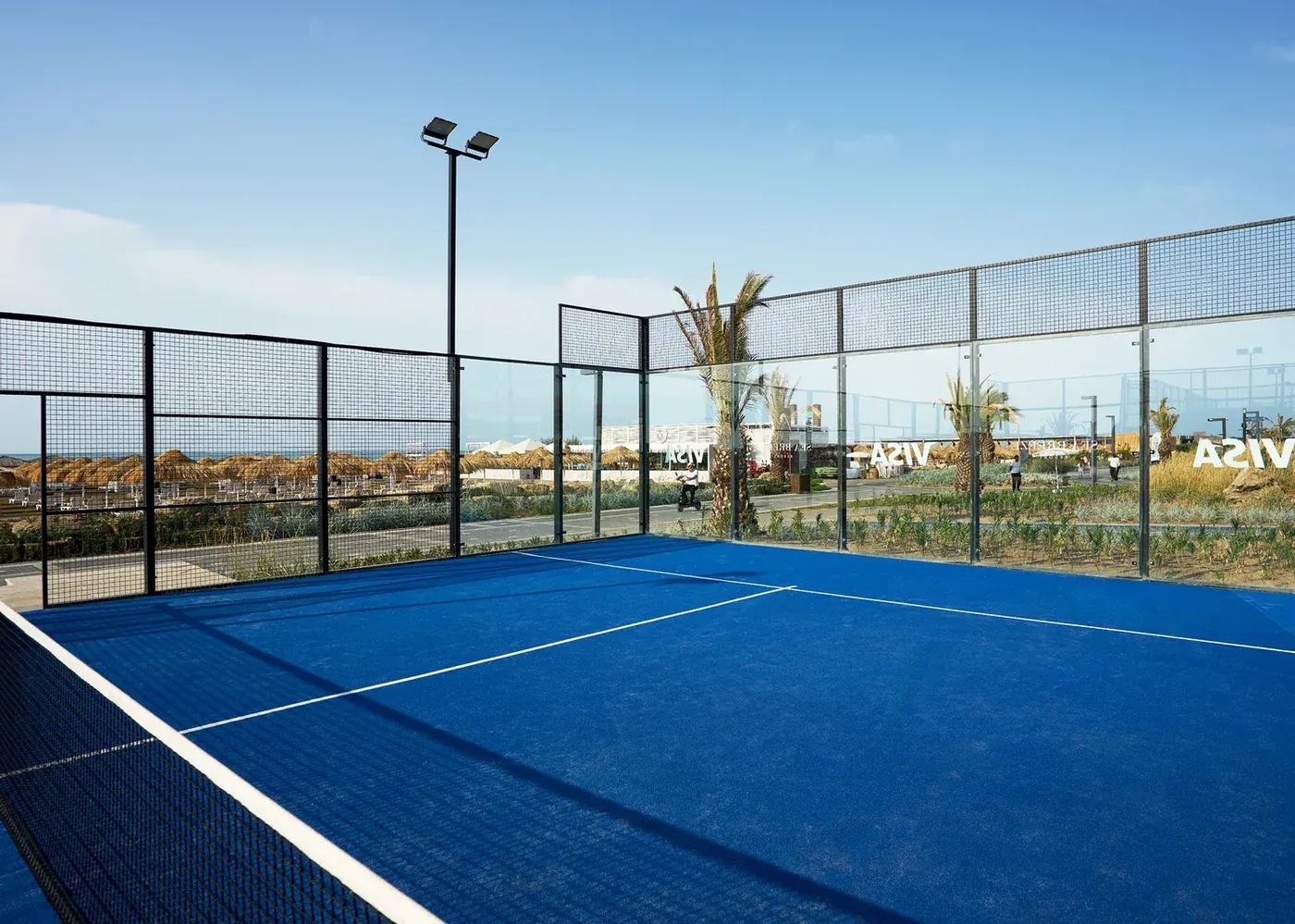 Şüşə bortlu mavi örtüklü padel kortu, işıqlandırma dirəkləri və palmalar